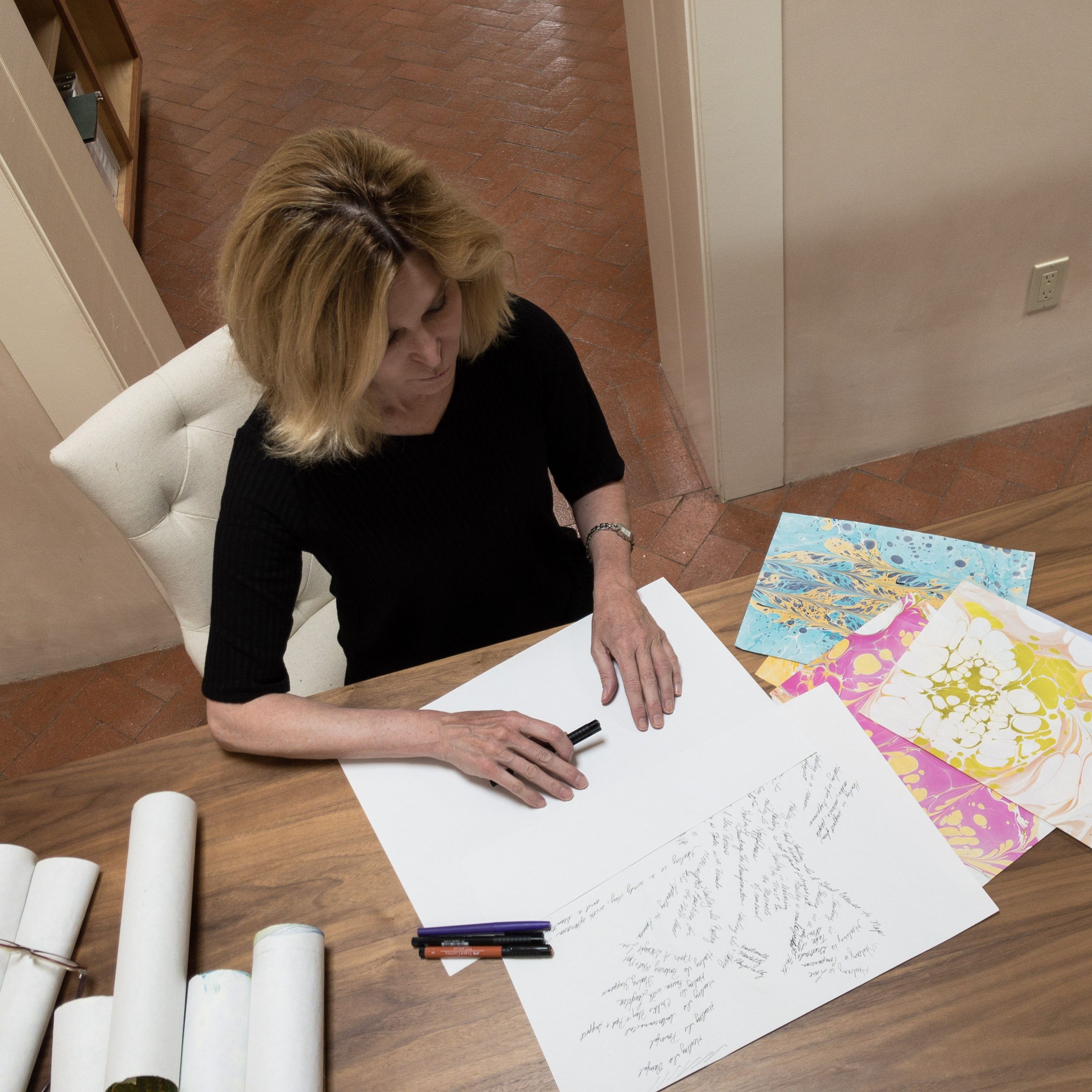 Patricia Varga sitting at a table with artwork and a blank sheet of paper