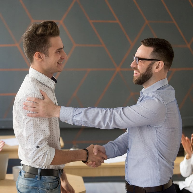 Two men in business casual shake hands while smiling