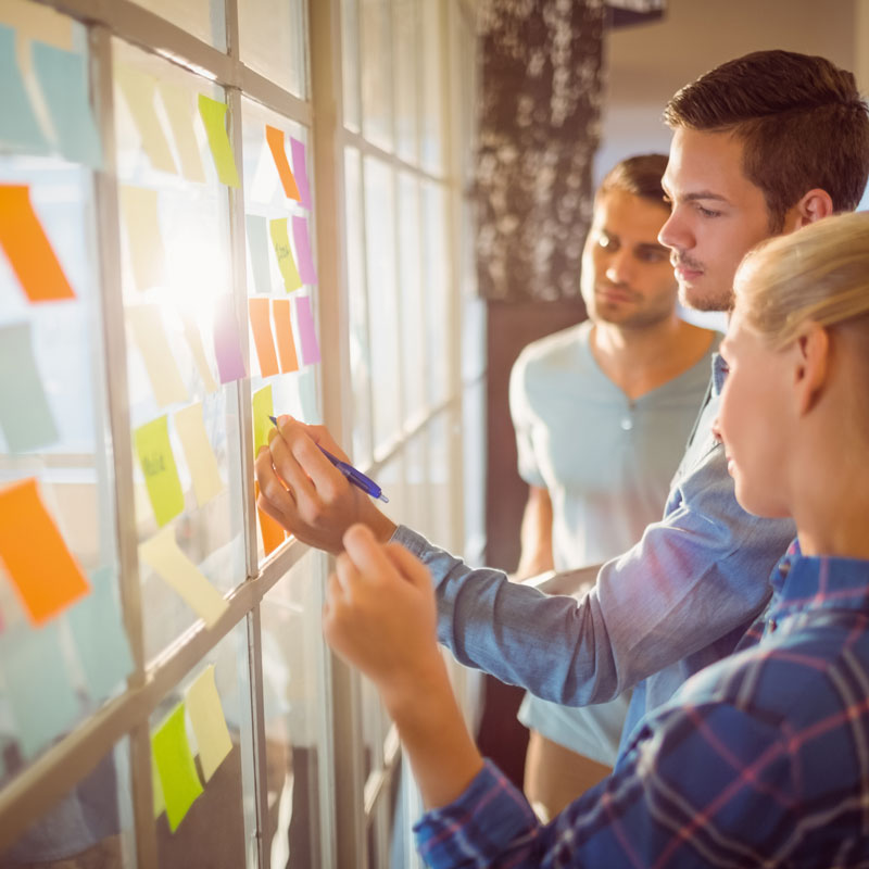 A team of one woman and two men stand by a glass window pane and write sticky notes